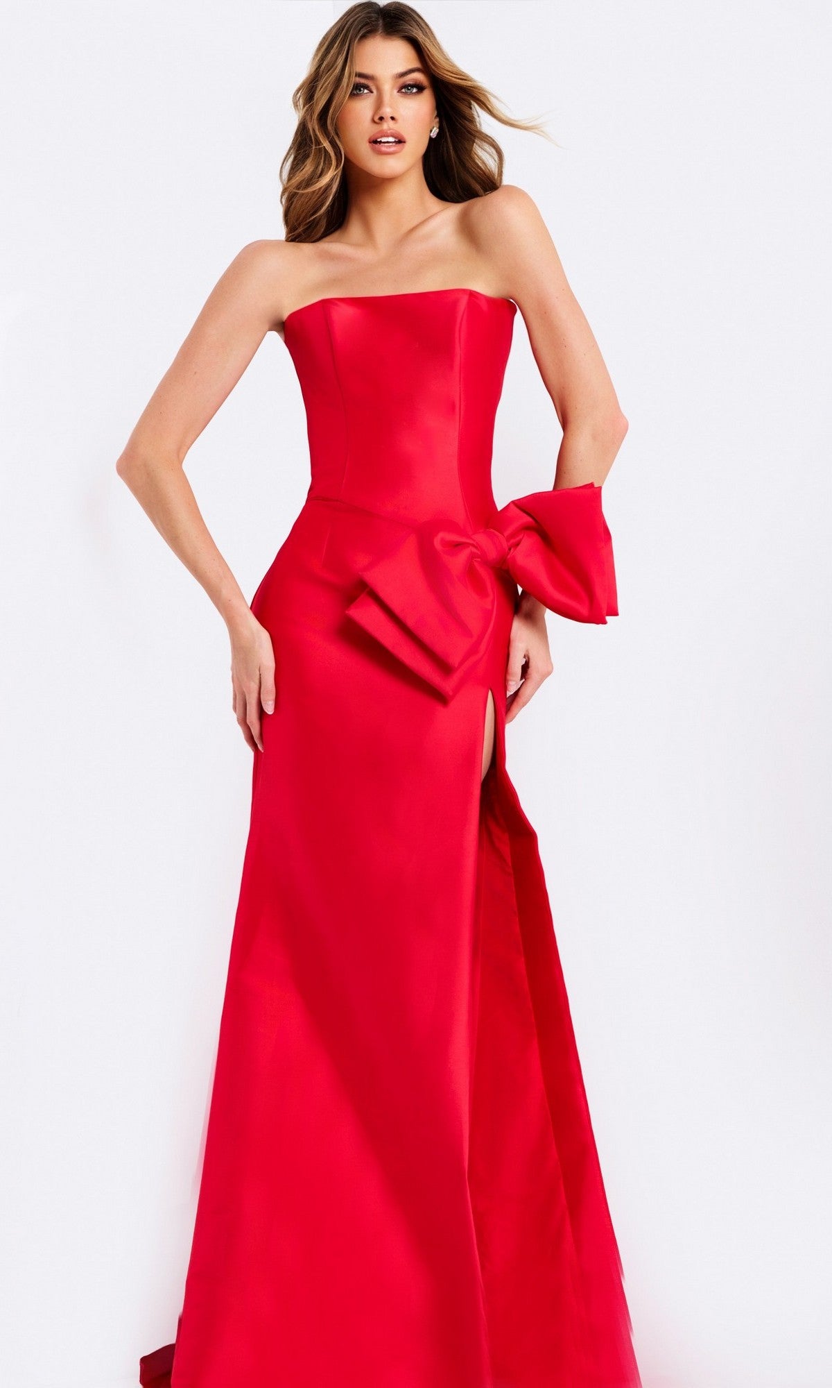 A woman stands against a white background in the Jovani 49195, a strapless, floor-length red satin prom dress featuring a large waist bow and thigh-high slit.